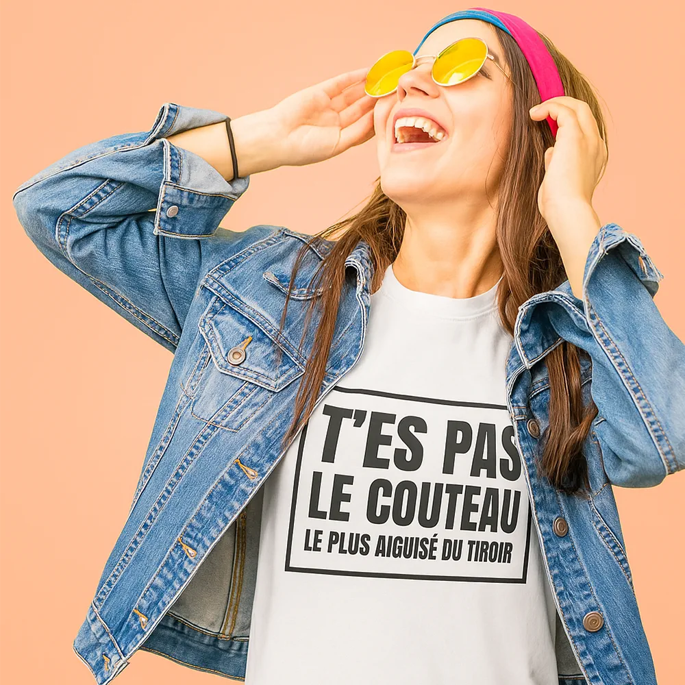 Femme souriante portant un t-shirt avec une phrase humoristique, vêtue d'une veste en jean et de lunettes de soleil, sur un fond de couleur unie.